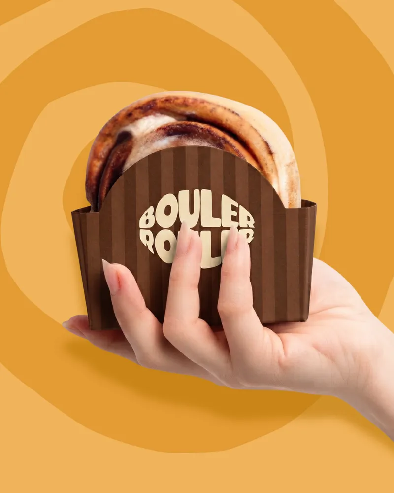 Une main tient un cinnamon roll dans un étui rayé brun portant le logo